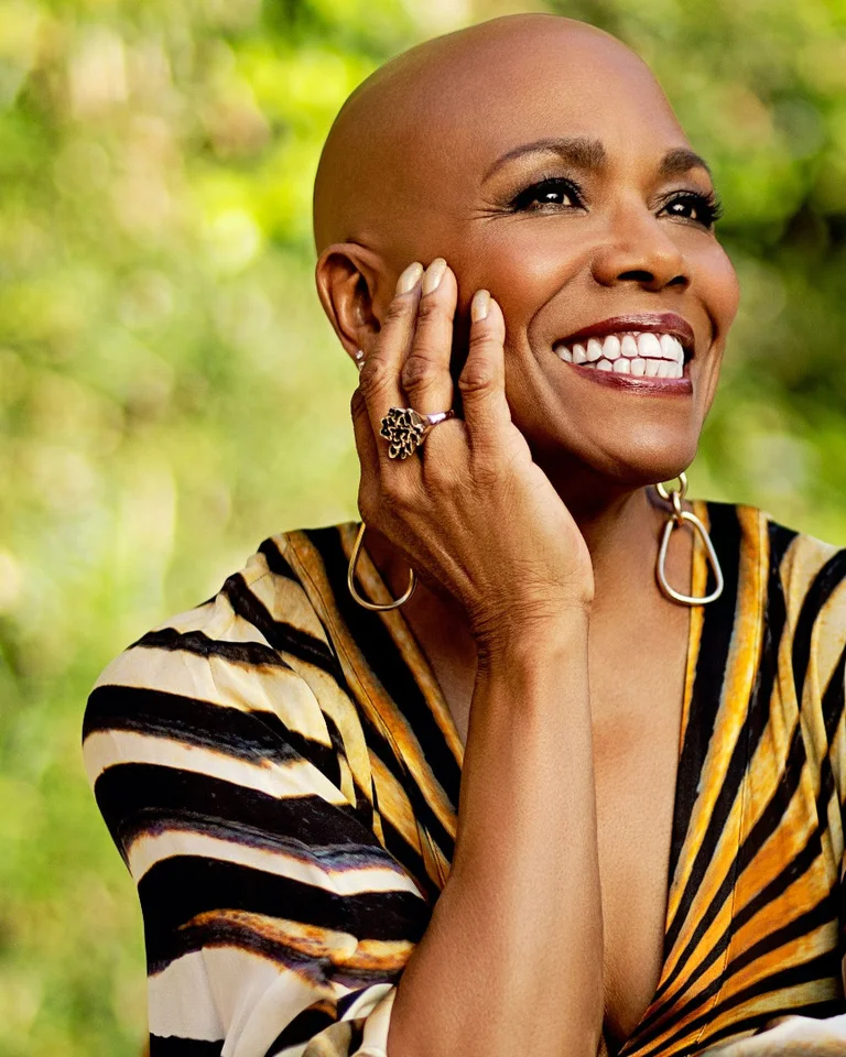 Eine lachende Person sitzt draußen, trägt ein gestreiftes Oberteil und stützt den Kopf auf die Hand. Hintergrund: grüne Pflanzen.