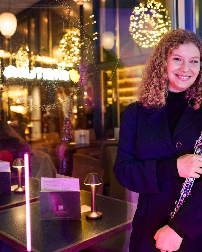 Eine Frau mit lockigem Haar, die eine Oboe hält, steht lächelnd vor einem elegant beleuchteten Café bei Nacht.