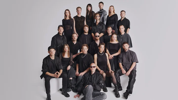 Group of 19 people wearing black clothes (Jazzrausch Bigband), posed against a gray background, arranged in tiered rows, some standing, some seated.