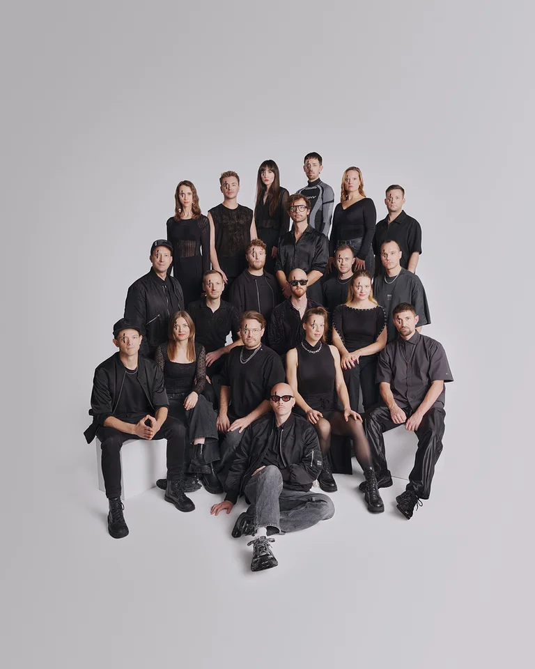 Group of 19 people wearing black clothes (Jazzrausch Bigband), posed against a gray background, arranged in tiered rows, some standing, some seated.