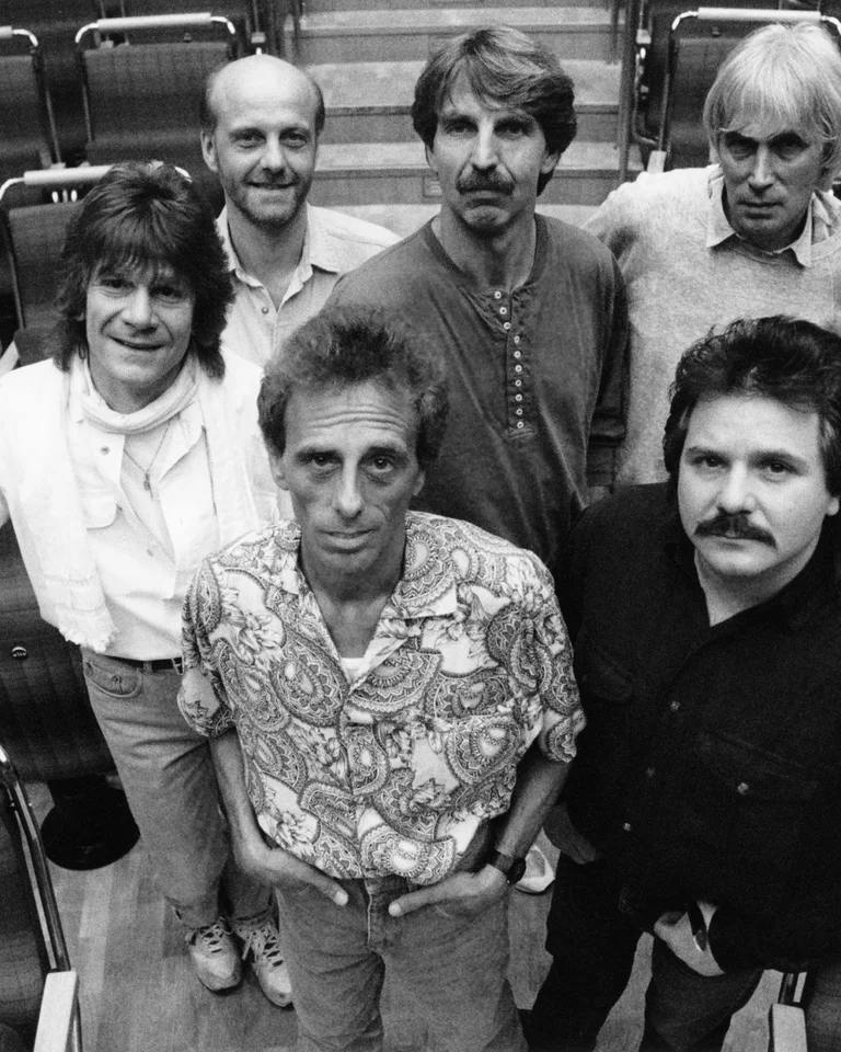 Black and white photo of six men standing in a theater-like space, looking towards the camera.