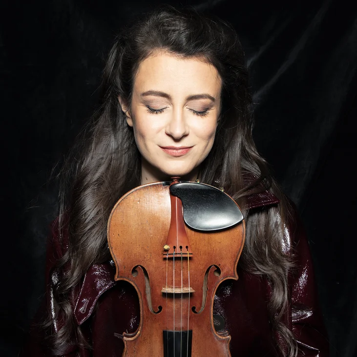 Eine Frau (Liya Petrova) steht mit geschlossenen Augen und hält eine Geige vor einem dunklen Hintergrund. Sie strahlt Ruhe aus.