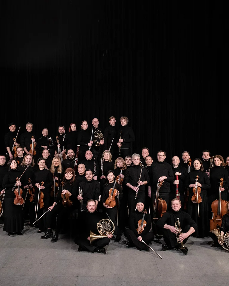 Ein großes Orchester mit Musikern in schwarzer Kleidung posiert mit verschiedenen Instrumenten vor einem dunklen Hintergrund.