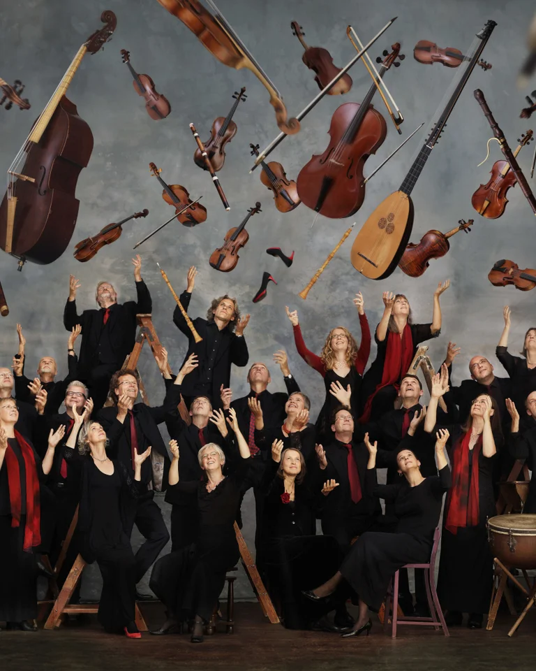 A group of people joyfully toss string instruments into the air. They wear black attire against a gray background.