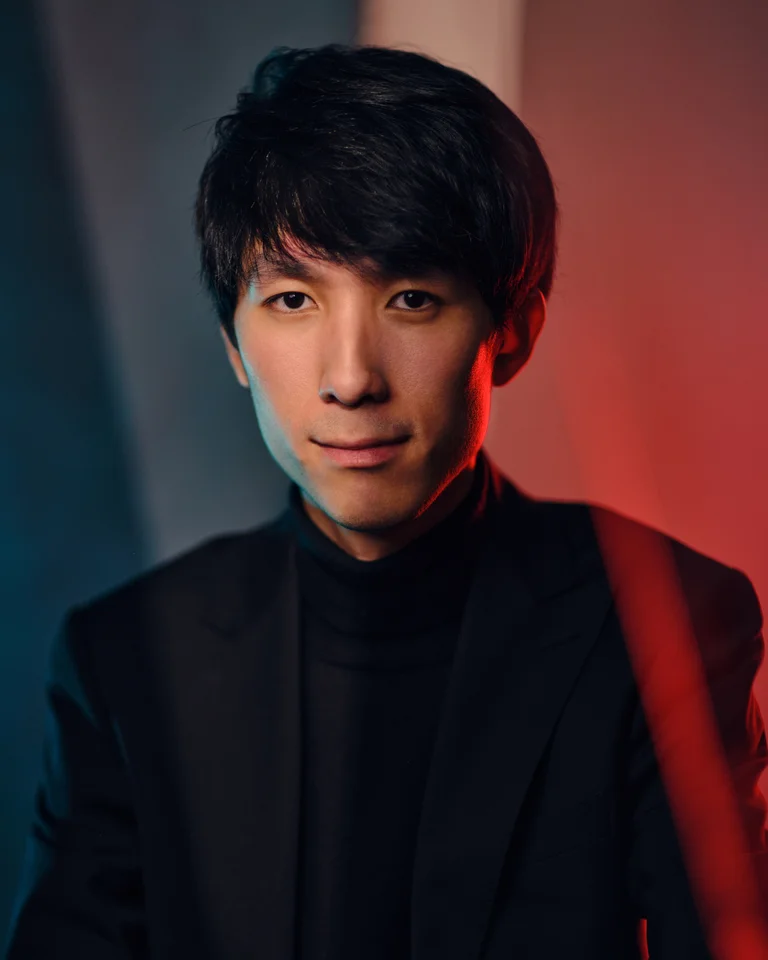 Porträt eines Mannes in einem dunklen Anzug vor einem rot-blauen Hintergrund, der freundlich in die Kamera blickt.