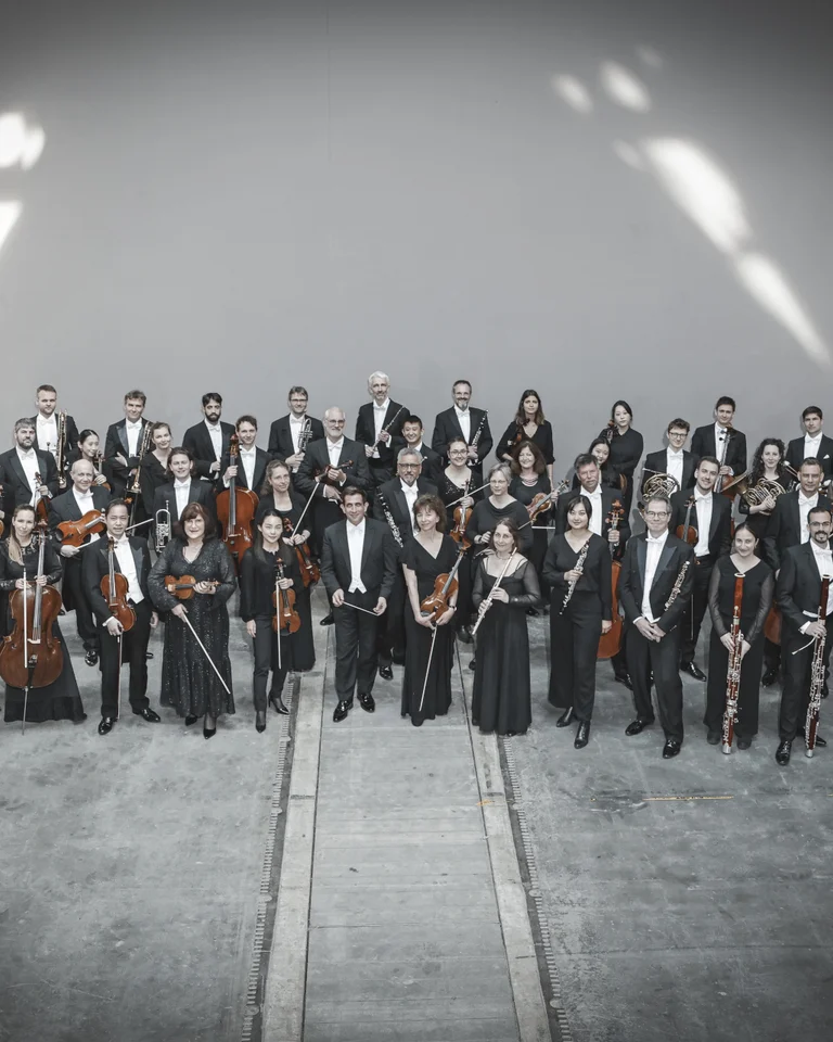 A large orchestra, dressed in formal evening attire, stands together in a spacious, sunlight-filled room with musical instruments.