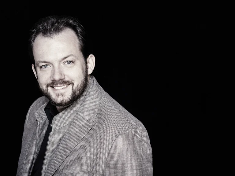 Andris Nelsons: Mann in grauem Anzug mit leichtem Lächeln vor schwarzem Hintergrund. Er hat Bart und kurzen Haarschnitt.