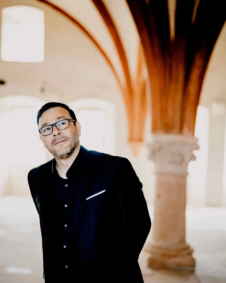 Ein Mann (Andreas Scholl) in einem eleganten, dunklen Anzug steht in einem Raum mit gotischen Bögen und historischer Architektur.