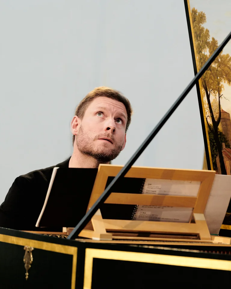 A man plays a harpsichord with forest painting in a serene room. He appears focused and contemplative.
