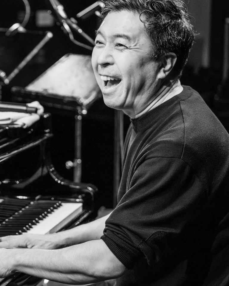 Ein Musiker spielt mit Freude Klavier auf einer Bühne. Noten und ein Mikrofon sind im Hintergrund sichtbar.