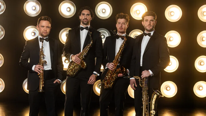 Vier Männer (SIGNUM Quartett) in Smokings stehen mit Saxophonen vor einer leuchtenden Hintergrundwand, die mit runden Lichtern dekoriert ist.
