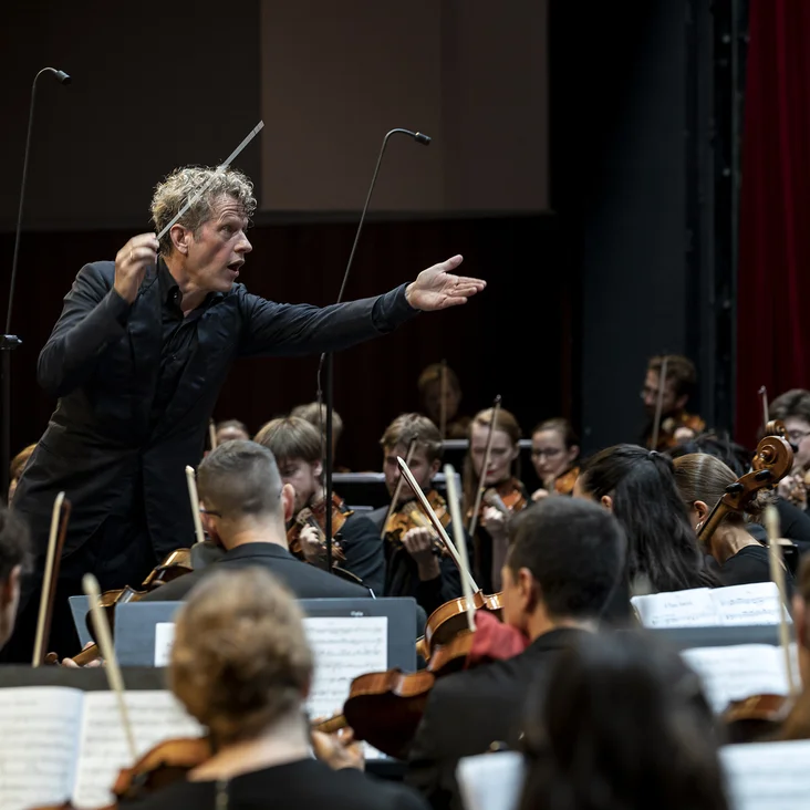 Ein Dirigent leitet ein großes Orchester (Philipp von Steinaecker und Mahler Academy Orchestra) während einer Aufführung. Die Musiker konzentrieren sich auf ihre Instrumente, umgeben von einem Konzertsaal.