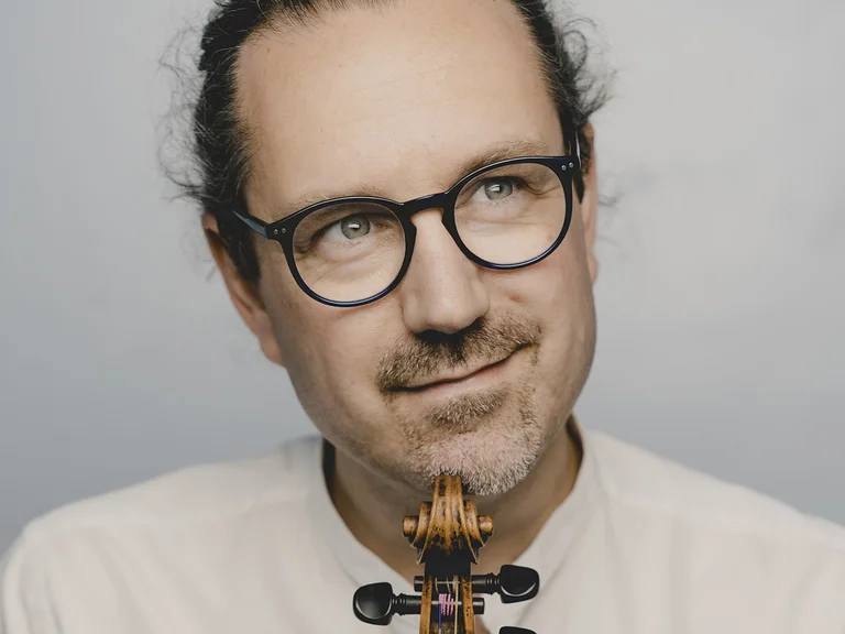 Porträt eines Mannes (Julien Chauvin) mit Brille und lockigem Haar. Er hält eine Geige und lächelt leicht.