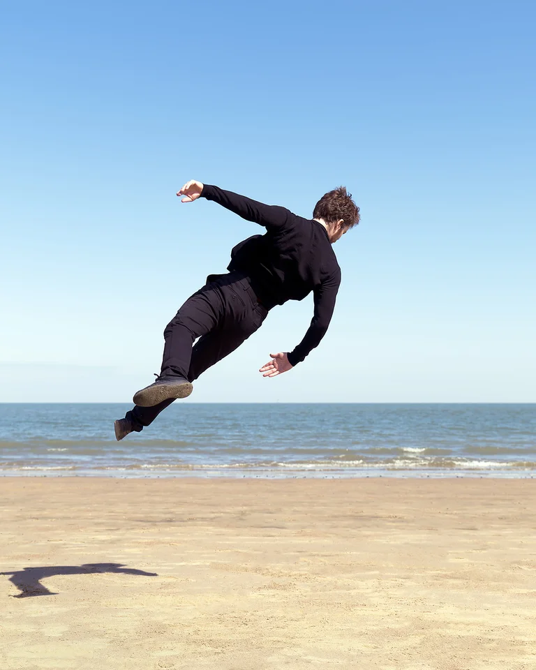 Eine Person in schwarzer Kleidung springt in die Luft über einem sandigen Strand mit dem blauen Horizont im Hintergrund.
