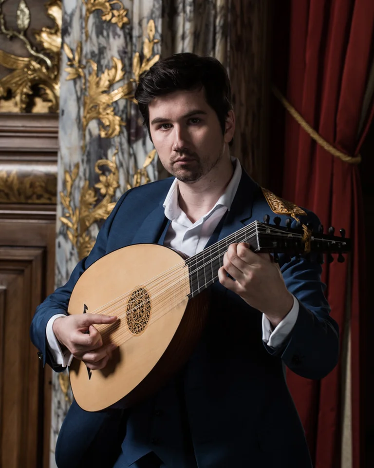 Ein Musiker (Thomas Dunford) in einem Anzug spielt eine Laute, umgeben von prächtigen Vorhängen und dekorativer Architektur im Hintergrund.