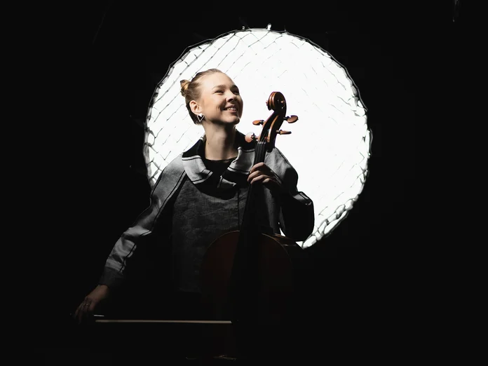 Valérie Fritz: Eine Cellistin, die vor einem hellen Lichtkreis spielt und lächelt. Der dunkle Hintergrund hebt ihre Silhouette hervor.