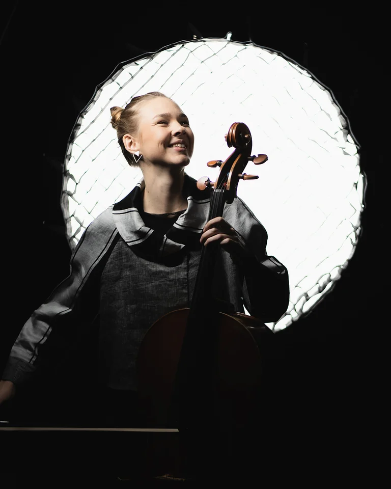 Valérie Fritz: Eine Cellistin, die vor einem hellen Lichtkreis spielt und lächelt. Der dunkle Hintergrund hebt ihre Silhouette hervor.