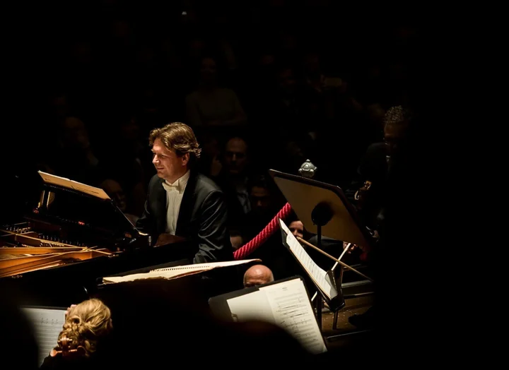 Ein Pianist in einem dunklen Konzertraum spielt konzentriert. Umgeben von Notenständern und Orchestermitgliedern, in gedämpftem Licht.