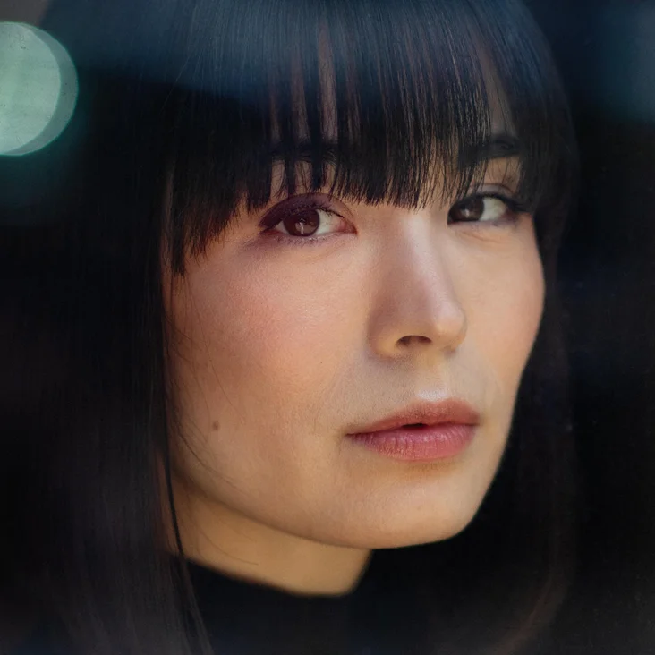 Portrait of a person with dark hair and bangs, gazing calmly at the camera. Blurred lights in the background.