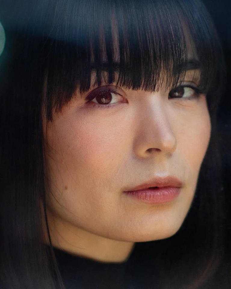 Portrait of a person with dark hair and bangs, gazing calmly at the camera. Blurred lights in the background.