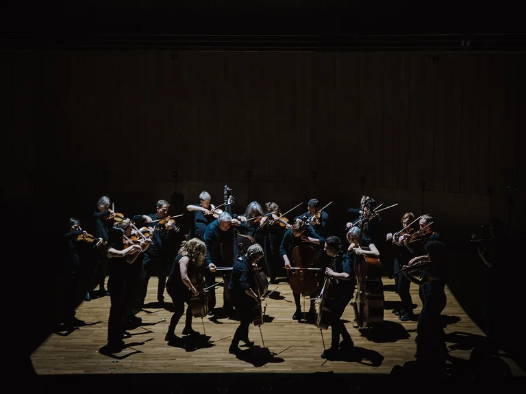 Ein Orchester (Scottish Ensemble) spielt auf einer spärlich beleuchteten Bühne mit Musikern, die Geigen und andere Streicherinstrumente spielen.