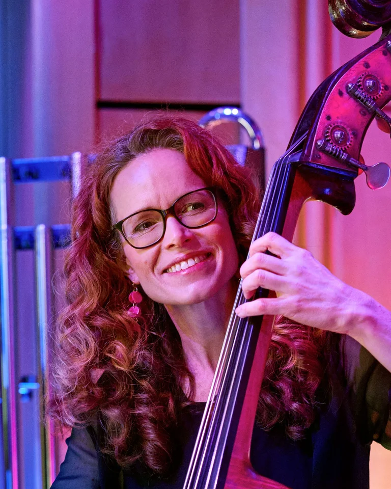 Eine Musikerin spielt einen Kontrabass auf einer Bühne, umgeben von farbigem Licht. Sie sieht zufrieden aus.