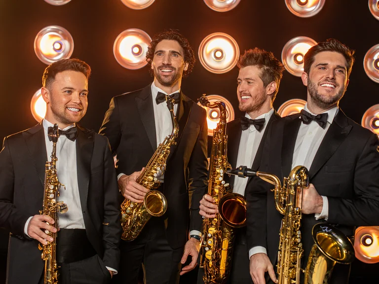 Vier Männer (SIGNUM Quartett) in Smoking halten Saxophone und lächeln. Sie stehen vor einer Wand aus runden Bühnenlichtern.