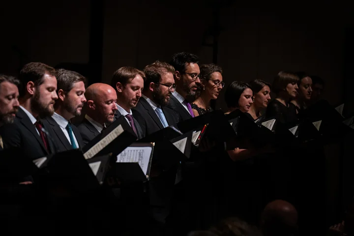 Members of the Choeur de chambre de Namur stand on a stage, all dressed in dark clothing. They are singing while holding sheets of music.