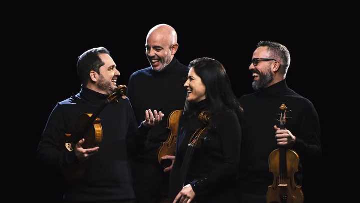 Vier Musiker (Cuarteto Quiroga) in schwarzer Kleidung stehen lächelnd mit ihren Streichinstrumenten vor einem dunklen Hintergrund und betrachten sich gegenseitig.