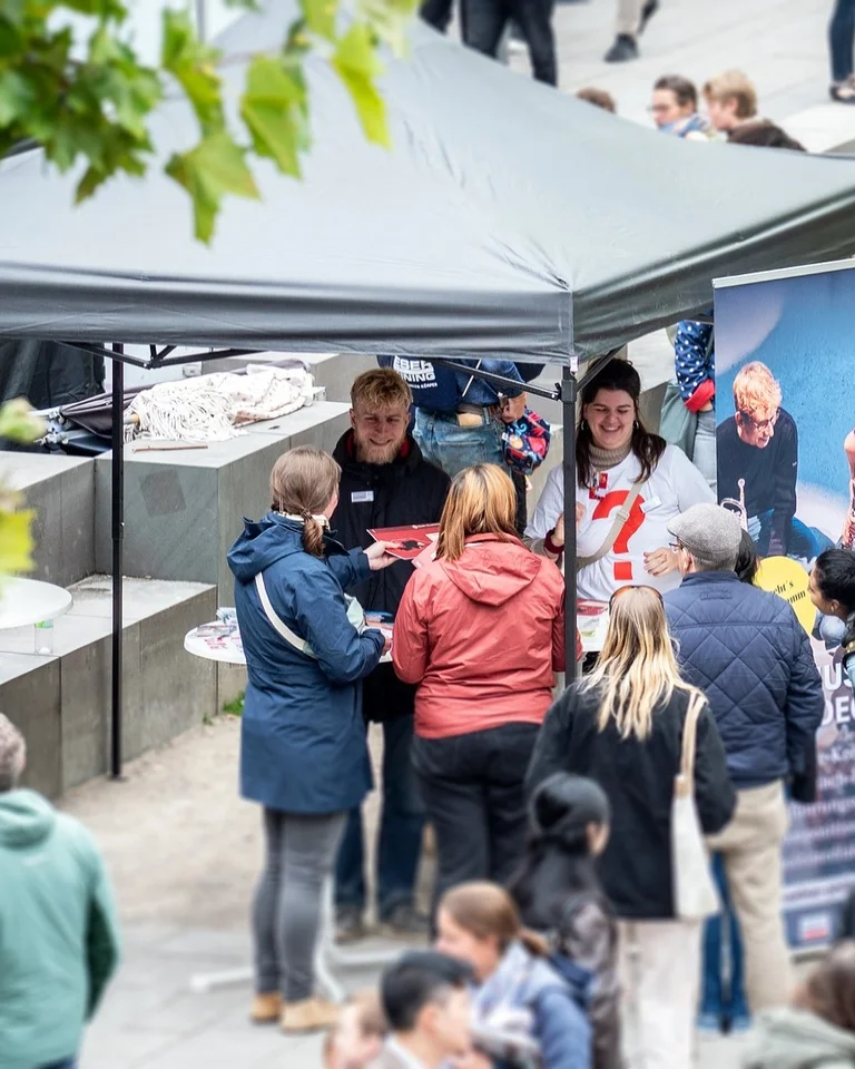 Menschen versammeln sich an einem Informationsstand unter einem Zelt. Einige sprechen mit den Standmitarbeitern, während andere in der Nähe verweilen.