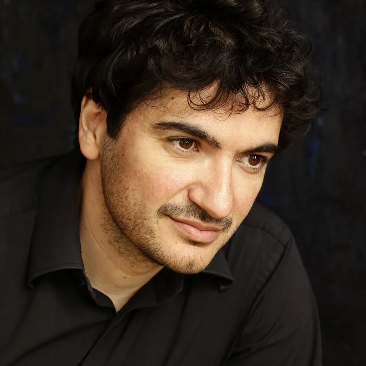 Portrait of Mathias Vidal with dark, curly hair and a beard, smiling in a friendly manner and wearing a dark shirt.