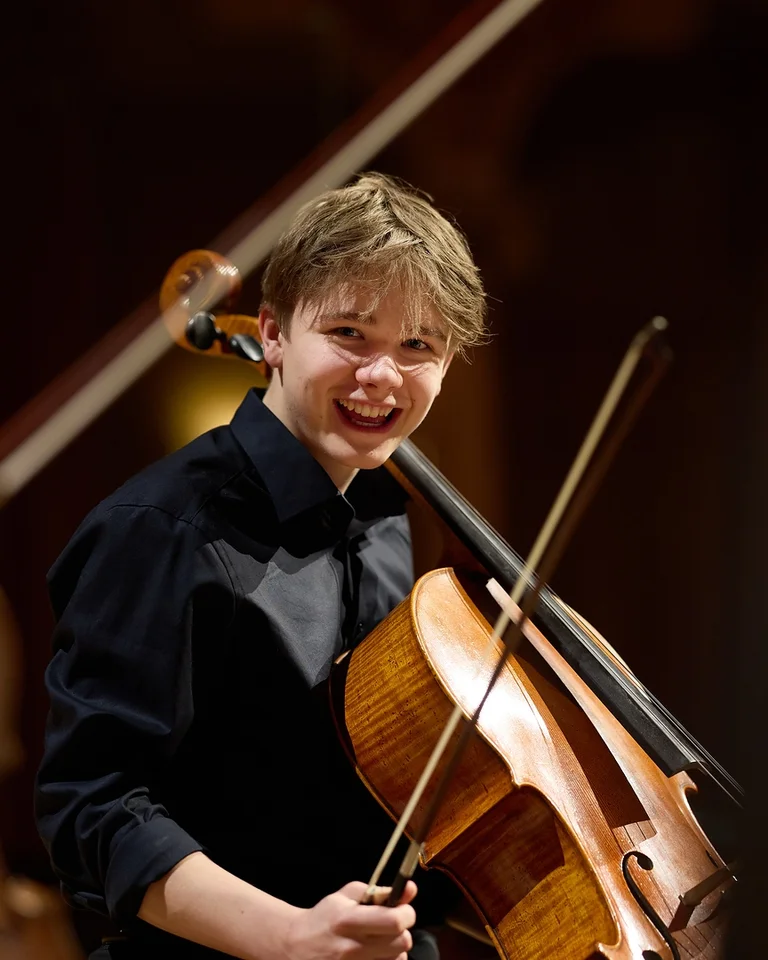 Ein junger Cellist lächelt während einer Aufführung auf der Bühne, neben einer Violinistin. Beide tragen Konzertkleidung.