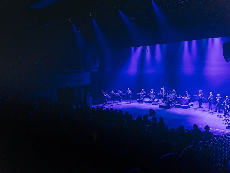 Eine Bühne in blauem Licht mit einer Reihe von Musikern (B'Rock), die vor einem Publikum orchestrale Instrumente spielen.