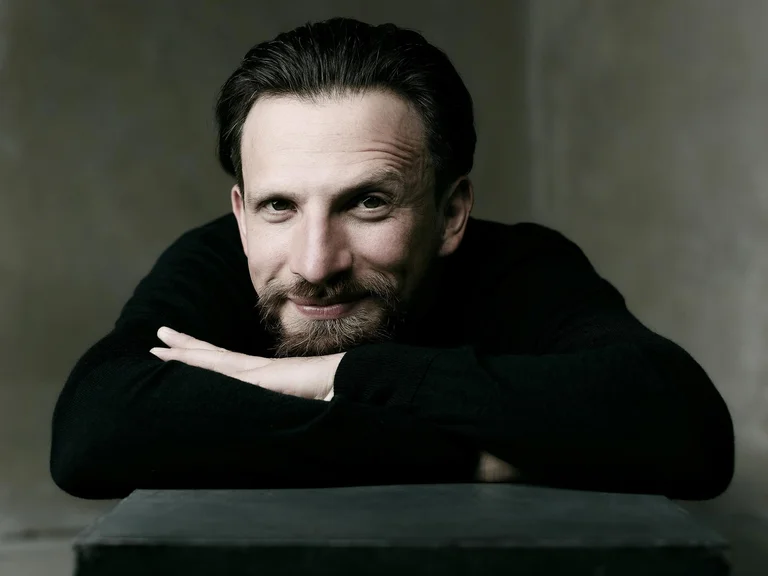 Ein lächelnder Mann (Cornelius Meister) lehnt sich auf eine schwarze Fläche und schaut direkt in die Kamera. Er trägt ein dunkles Oberteil.