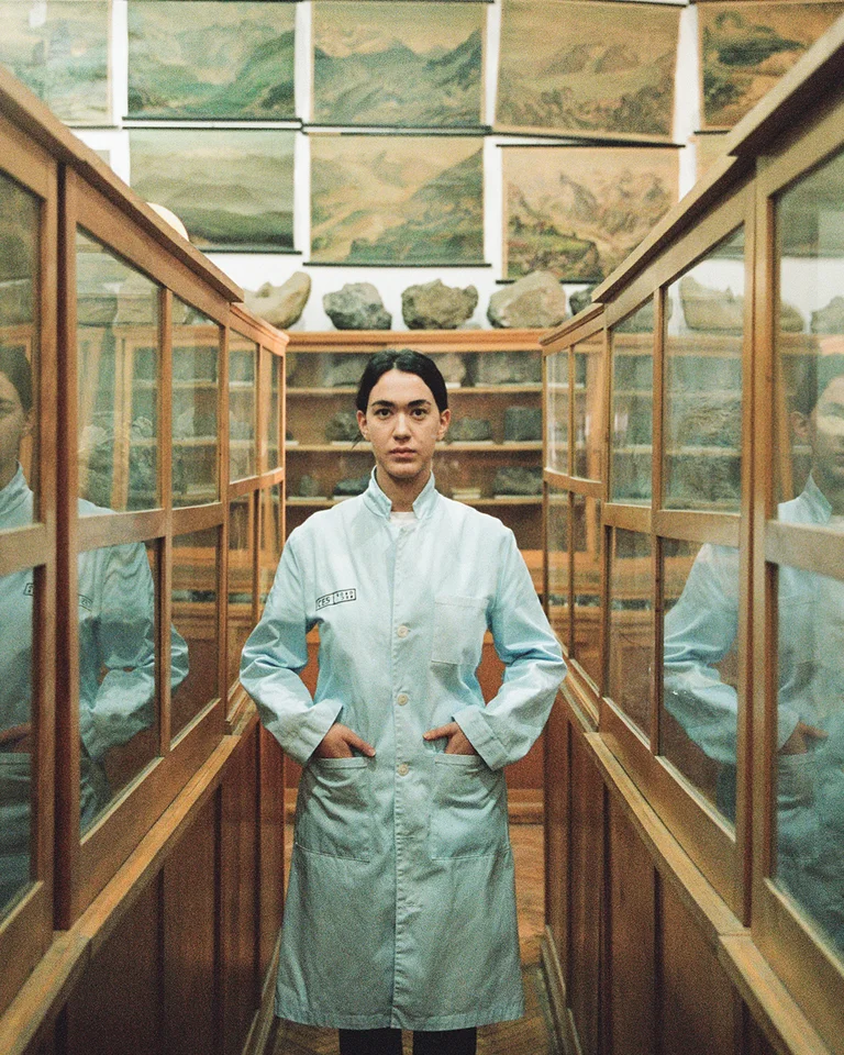 Person (Anushka Chkheidze) in weißem Laborkittel steht zwischen Glasvitrinen in einem antik wirkenden Raum mit regalen. Beide Seiten sind voller Artefakte.