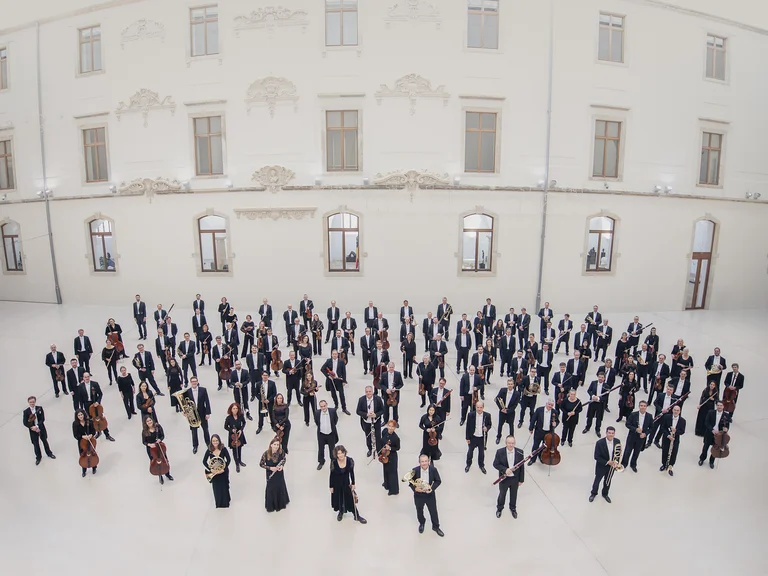 Eine große Gruppe von Orchestermusikern (Sächsische Staatskapelle Dresden) steht draußen vor einem historischen Gebäude. Sie tragen formelle Kleidung und halten Instrumente.