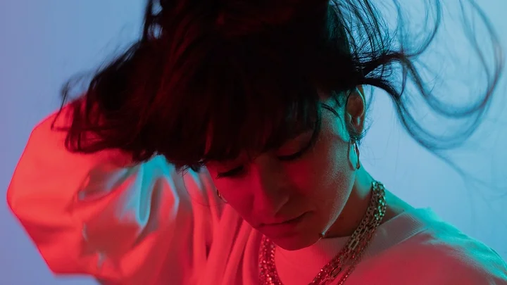 Eine Person mit geschlossenen Augen schüttelt ihr Haar unter blau-roter Beleuchtung. Sie trägt ein weißes Oberteil und Halsketten.