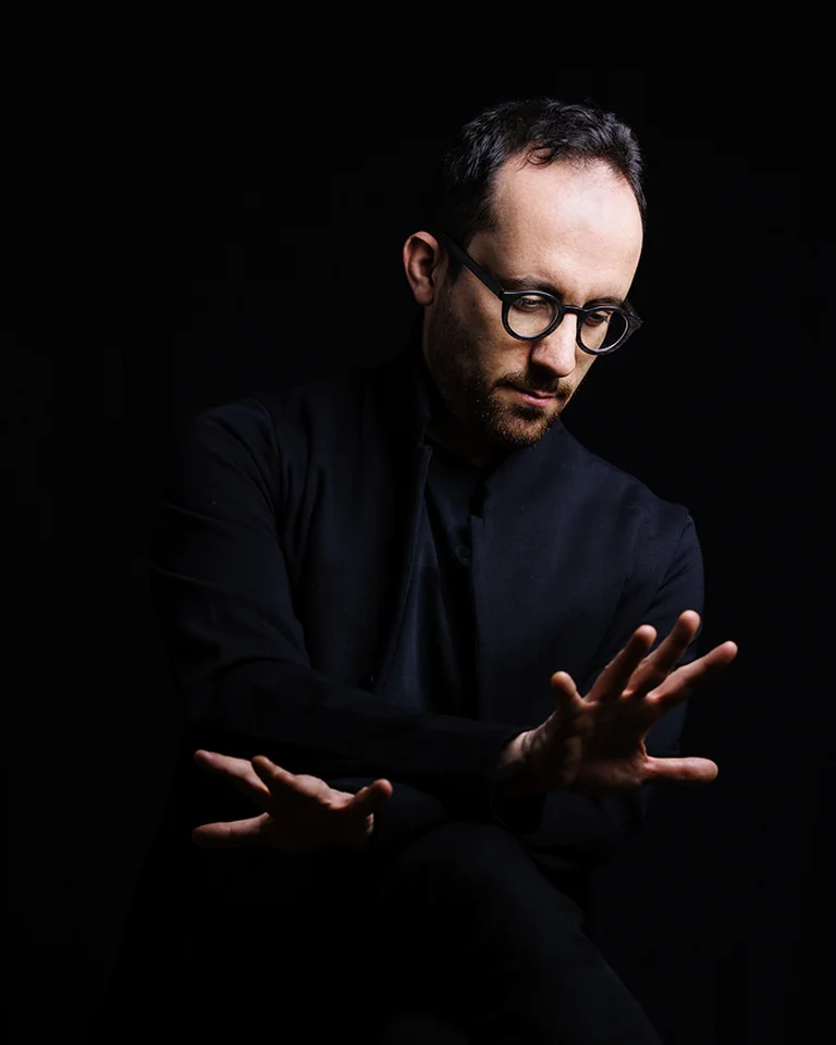 Ein Mann in schwarz gekleidet (Igor Levit) sitzt vor einem dunklen Hintergrund. Er trägt eine Brille und gestikuliert mit seinen Händen.