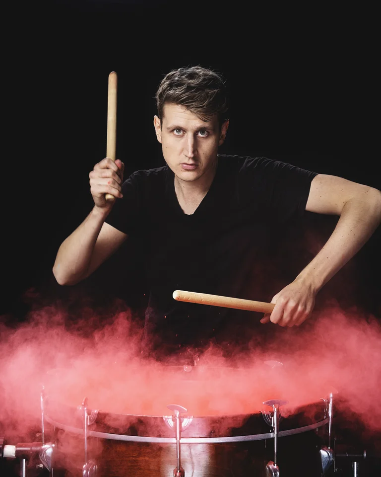 A drummer energetically plays a drum with red clouds of powder rising. The background is dark, highlighting the performer.
