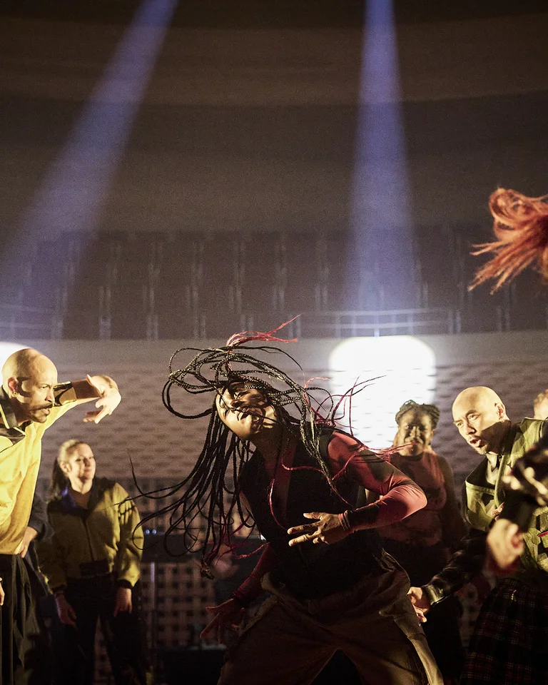 Dancers in energetic poses on stage with dramatic lighting. They wear creative outfits, showcasing movement and expression.