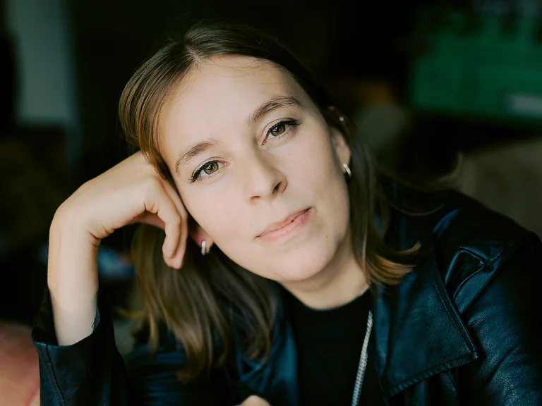 Eine Frau (Charlotte Oelschlegel) mit brauner Jacke stützt ihren Kopf auf ihre Hand, sitzt auf einem gepolsterten Sofa und blickt entspannt.