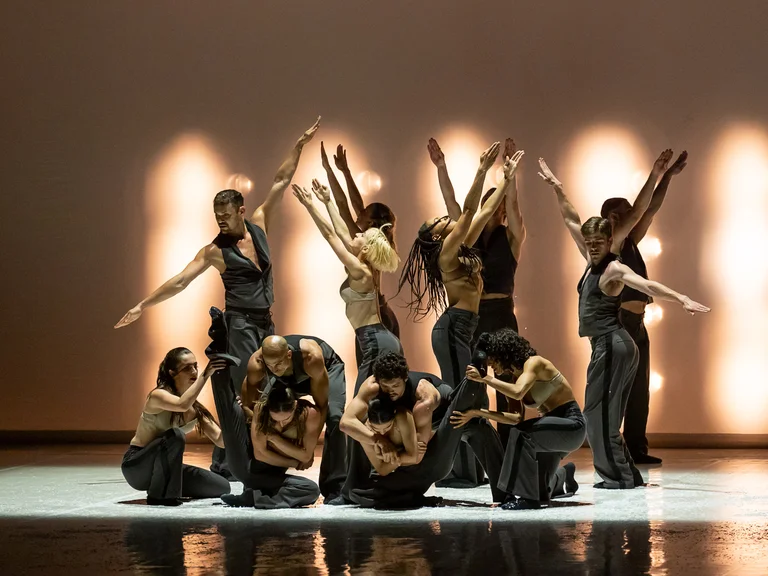 Tänzer auf einer Bühne in dynamischen Posen vor einem beleuchteten Hintergrund. Sie tragen dunkle Kleidung und bewegen sich synchron.