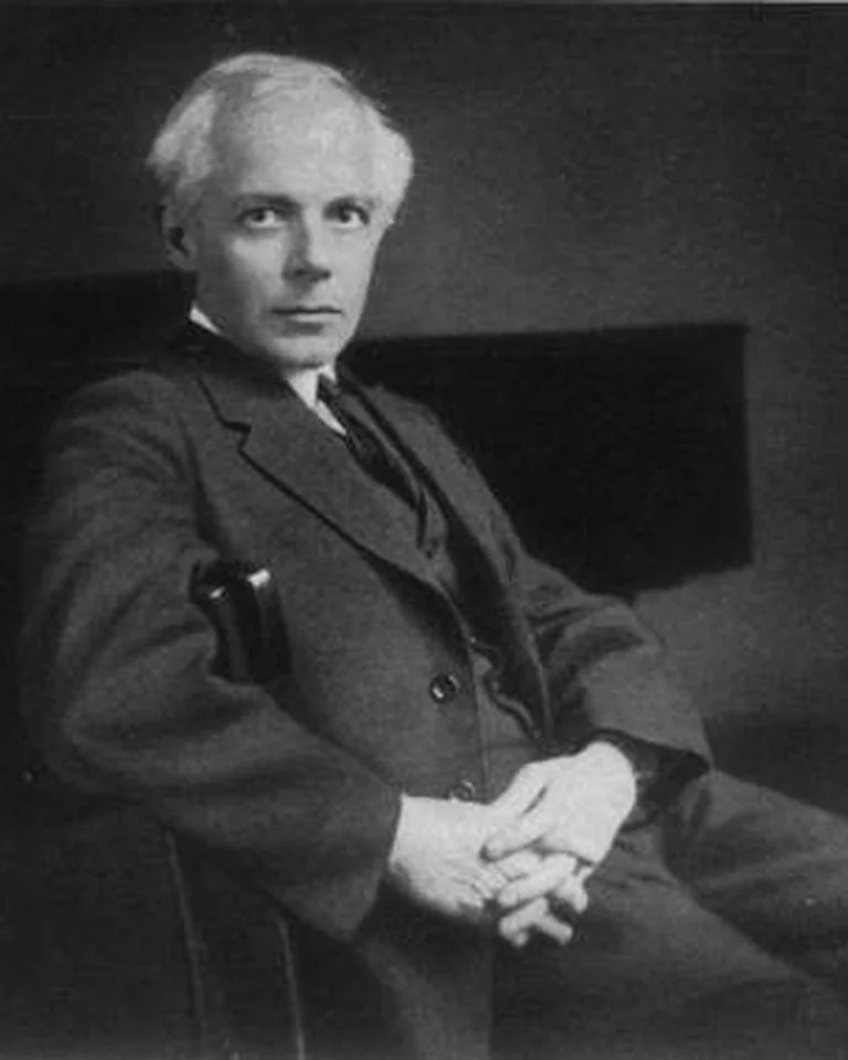 Ein älterer Mann in einem Anzug (Béla Bartók) sitzt entspannt auf einem Stuhl. Der Hintergrund ist dunkel und unscharf.