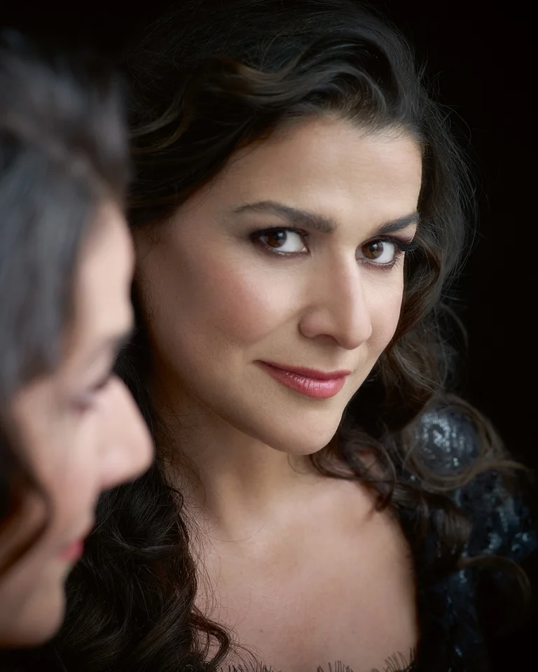 Porträt einer Frau mit langem, dunklem Haar, die leicht lächelt. Ihr Bild spiegelt sich im Hintergrund wider.