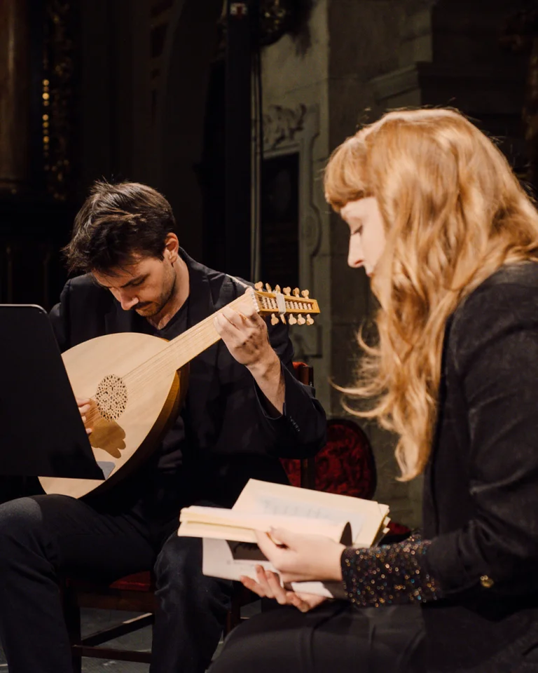 Eine Frau liest aus einem Buch, während ein Mann Laute spielt. Beide sitzen auf Stühlen in einem dunklen Raum.