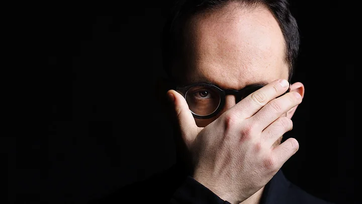 Eine Person in dunkler Kleidung (Igor Levit) bedeckt teilweise ihr Gesicht mit der Hand, vor einem schwarzen Hintergrund. Sie trägt eine Brille.