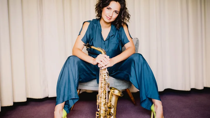 Eine Frau sitzt lässig auf einem Sessel und hält ein Saxofon. Sie trägt einen blauen Anzug und gelbe Absätze.