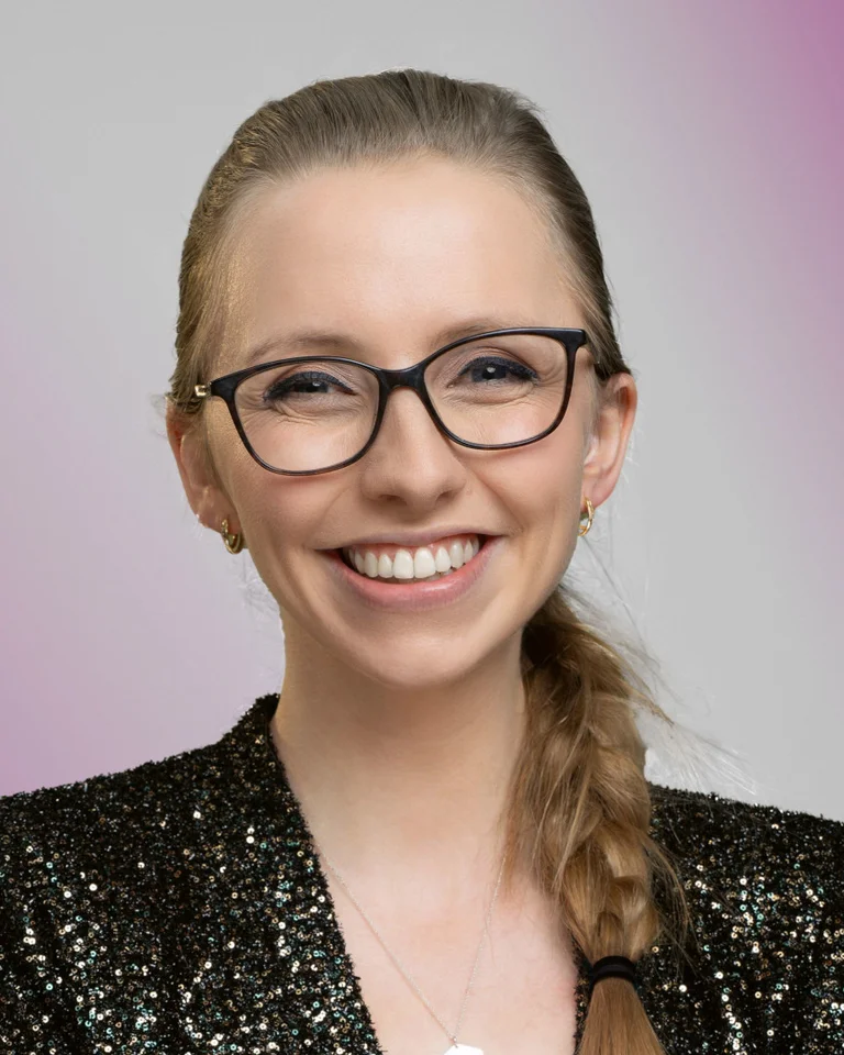Die Person lächelt und trägt eine Brille und einen schwarzen Blazer mit Glitzer-Effekt. Der Hintergrund ist lila.