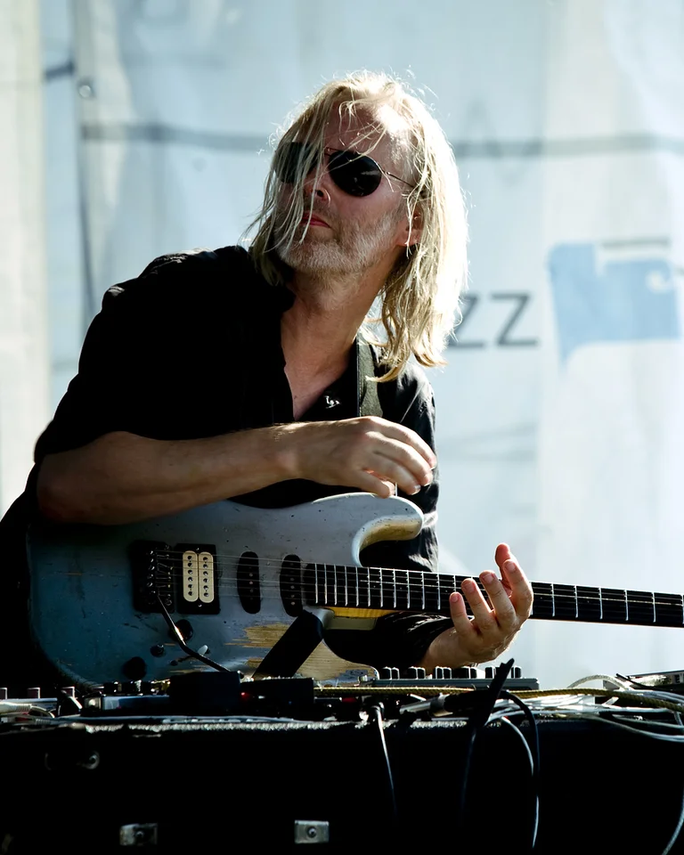 A musician with sunglasses plays a guitar in front of a laptop. The background features graffiti and a jazz banner.