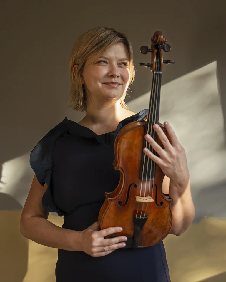 Eine lächelnde Frau (Alina Ibragimova) in einem dunklen Kleid hält eine Geige. Weiches Licht fällt auf sie und die neutrale Wand.